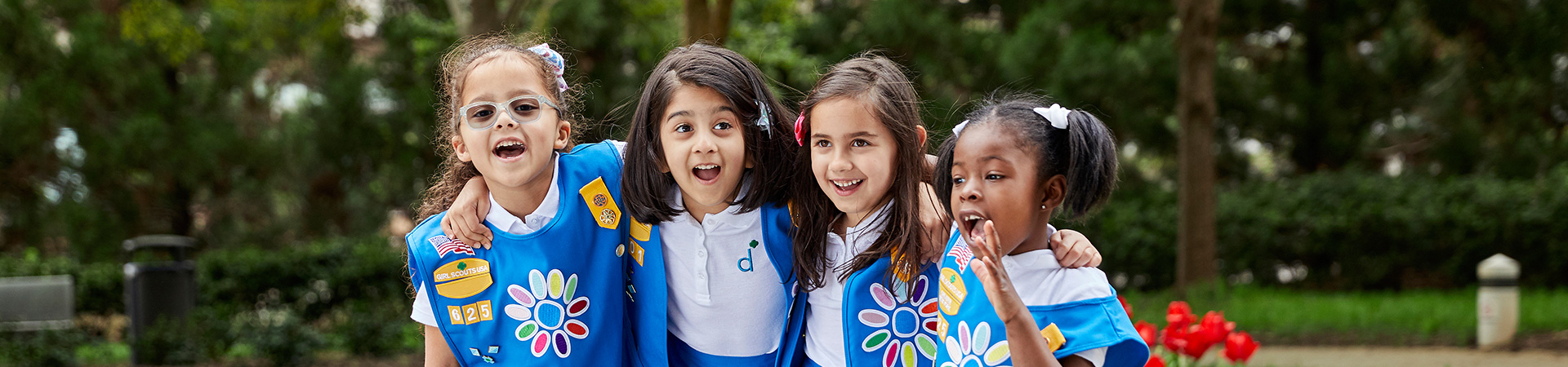  Group of kindergarten daisy girl scouts in vest and apron uniform hugging and smiling outside. 